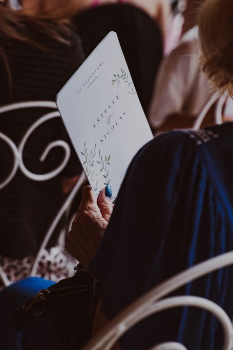 Une main tient un programme imprimé lors d'un événement, avec des personnes en arrière-plan.