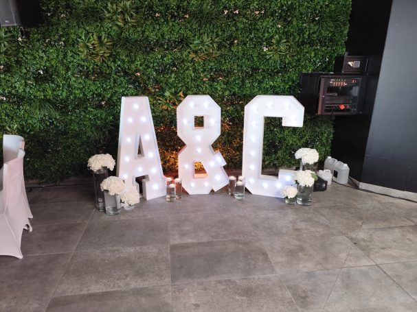 Lettres lumineuses "A" et "C" décorées de fleurs, devant un mur de verdure.