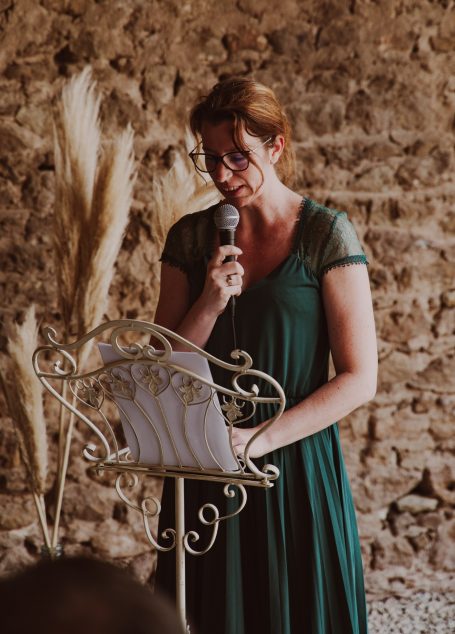 Femme en robe verte souriant et tenant un micro, parlant devant un pupitre décoratif.