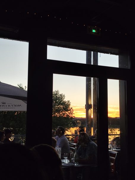 Vue sur un coucher de soleil à travers une fenêtre, avec des silhouettes de personnes à l'intérieur.