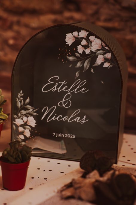 Panneau en verre avec les noms Estelle et Nicolas, décoré de fleurs blanches.