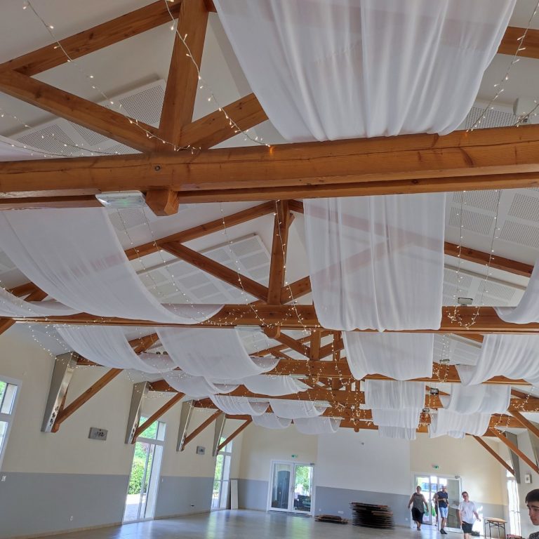 Plafond d'une salle avec poutres en bois et voiles blancs, ambiance lumineuse et aérée.